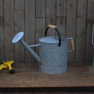 Photo d'un arrosoir vintage en zinc gris à poignées en bois une anse fixe et une mobile noire, placé sur une planche de jardin devant une palissade en bois un objet de jardin jaune posé à sa gauche