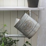 Photo d'un arrosoir vintage gris suspendu au crochet d'une étagère par l'anse arrière carrée une impression magnolia greenhouse nursery au centre devant une palissade en bois gris clair et une plante sur la gauche