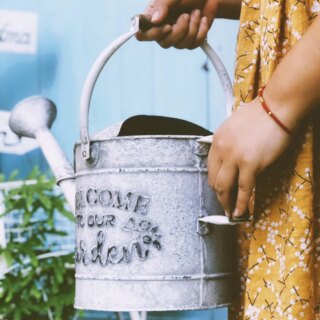Photo d'un arrosoir vintage gris style ferme avec deux poignées en bois et inscription Welcome to our garden tenu par une femme en robe jaune moutarde qui s'apprête à arroser une plante le tout sur fond bleu clair