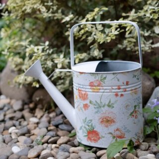 Photo d'un arrosoir vintage blanc à fleurs aux tons doux orange saumon et vert d'eau à anse carrée posé sur un chemin de cailloux devant un massif de plantes