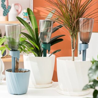Photo représentant 2 pots de plantes blancs et un bleu plus petit munis de système d'arrosage goutte-à-goutte en fonction posés sur une table blanche avec un mur orange en fond